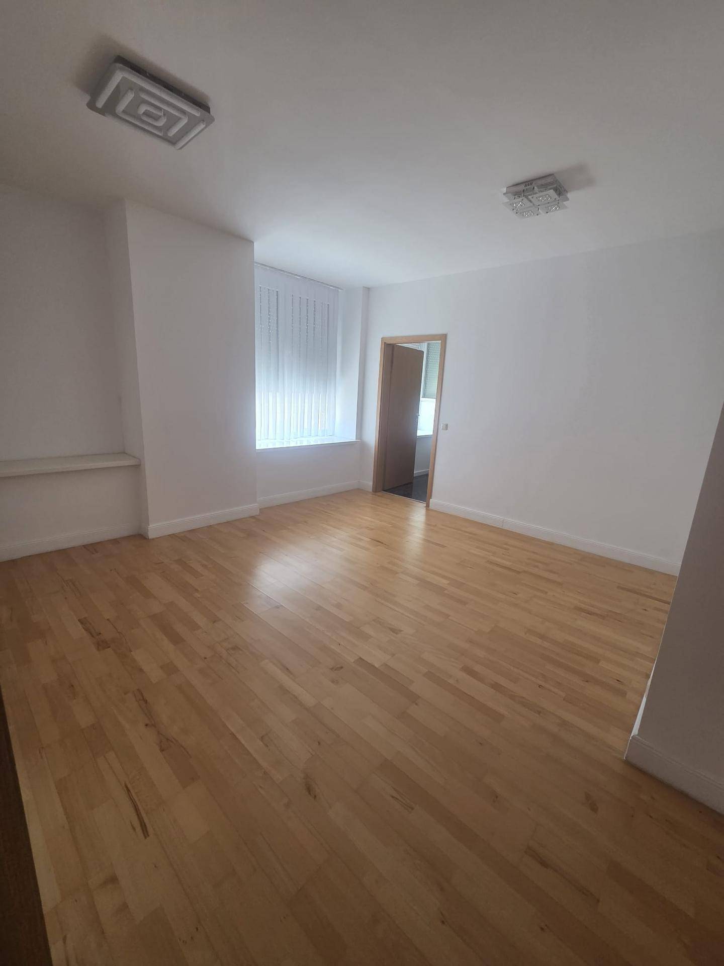 Helles Wohnzimmer mit Holzboden, weißen Wänden und einem Fenster mit Jalousien.