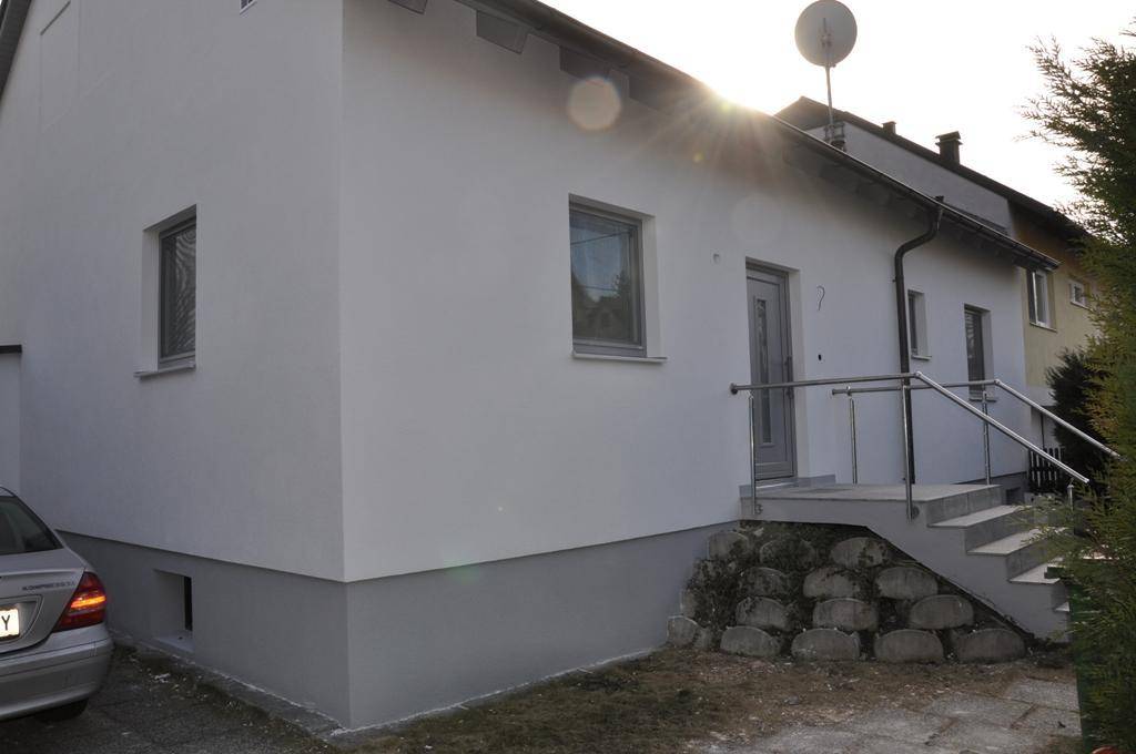 Modernes Einfamilienhaus mit weißer Fassade und grauem Sockel, sonniger Eingangsbereich mit Treppe.