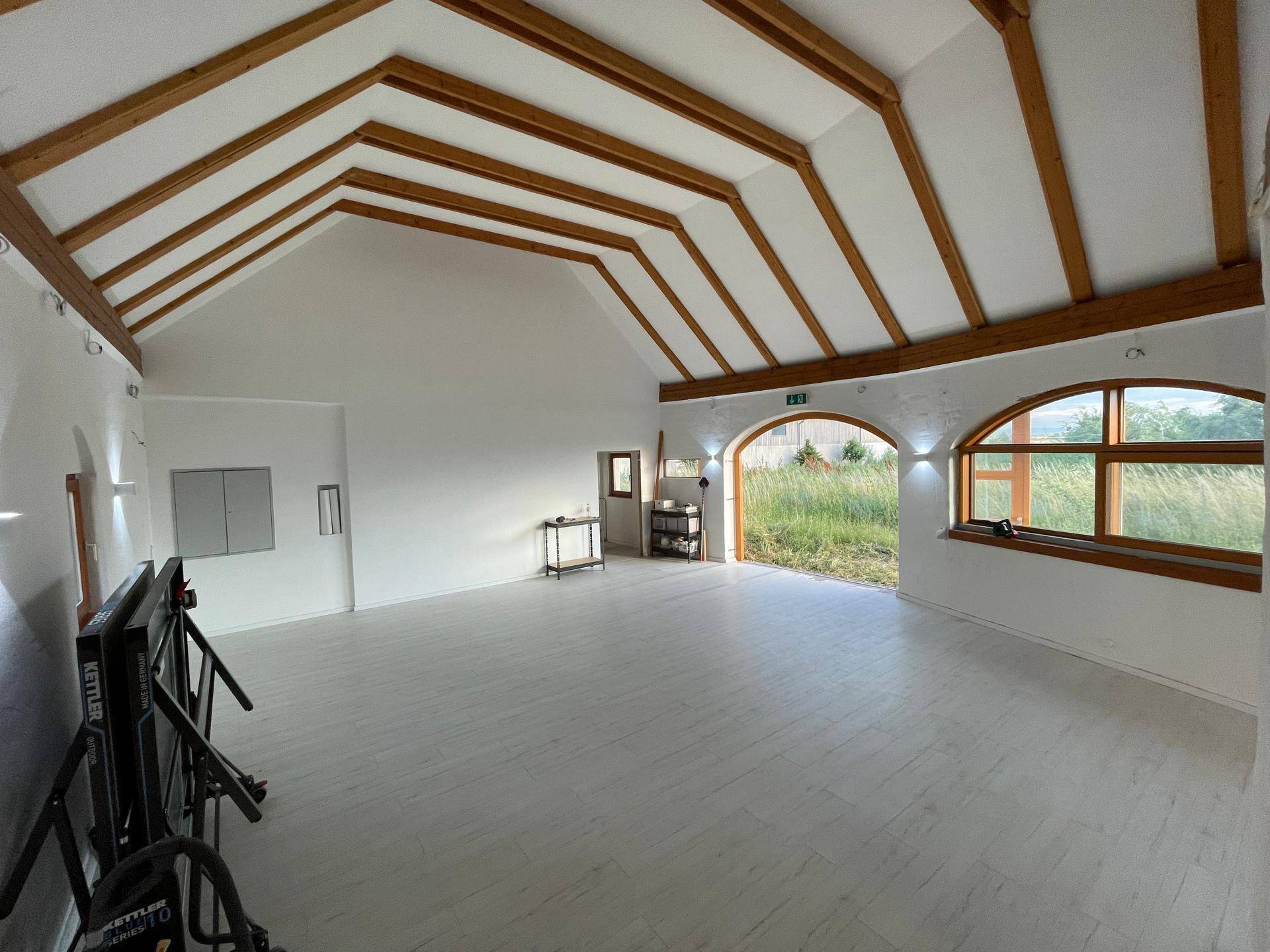 Geräumiger Innenraum mit hohen Decken, sichtbaren Holzbalken und hellem Boden, ideal für vielseitige Nutzung.
