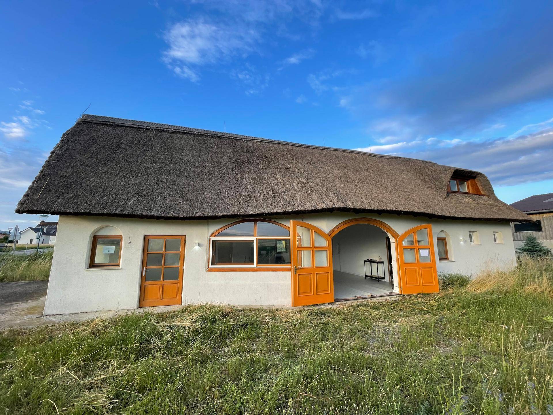 Reetgedecktes Haus mit geöffneten Holztüren, die einen Einblick in den Innenraum gewähren, inmitten einer Naturlandschaft.