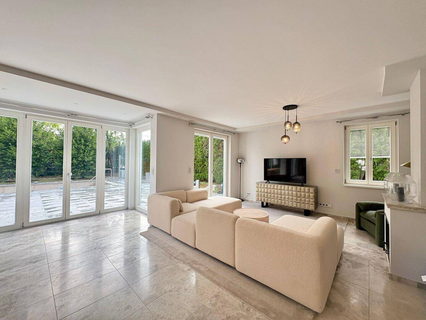 Geräumiges Wohnzimmer mit großen Fenstern, modernem Sofa und hellem Fliesenboden.