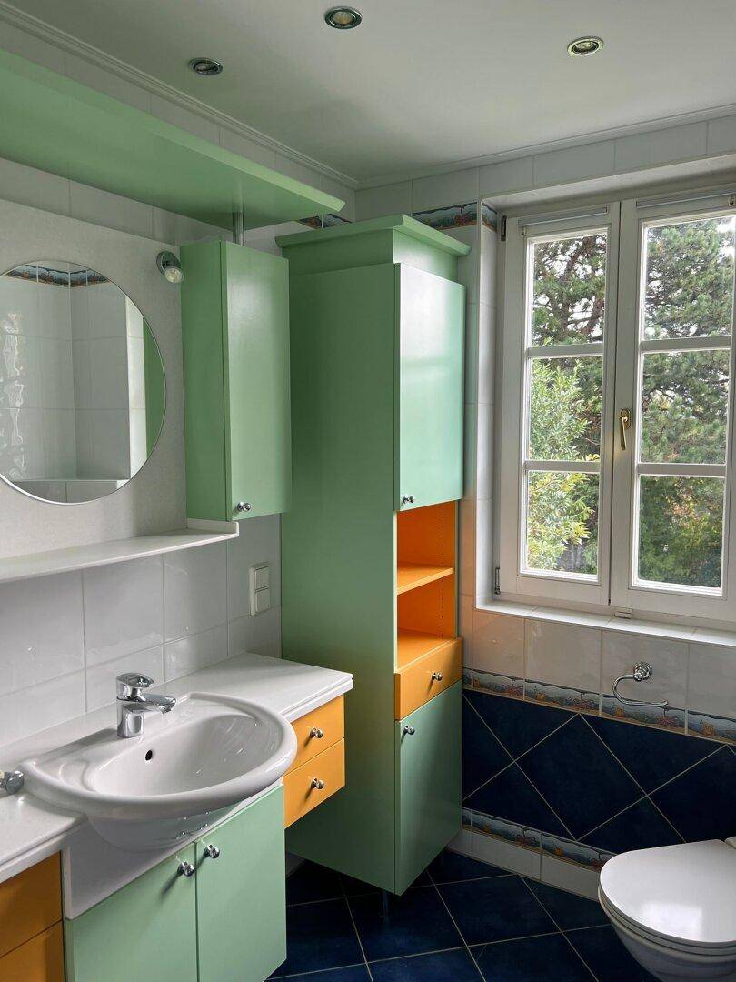Farbiges Badezimmer mit Fenster, Waschtisch und auffälligen grünen und orangen Schränken.