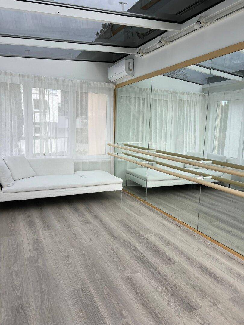 Heller Fitnessraum mit Spiegelwand, Ballettstangen, Sofa und Klimaanlage unter Glasdach.