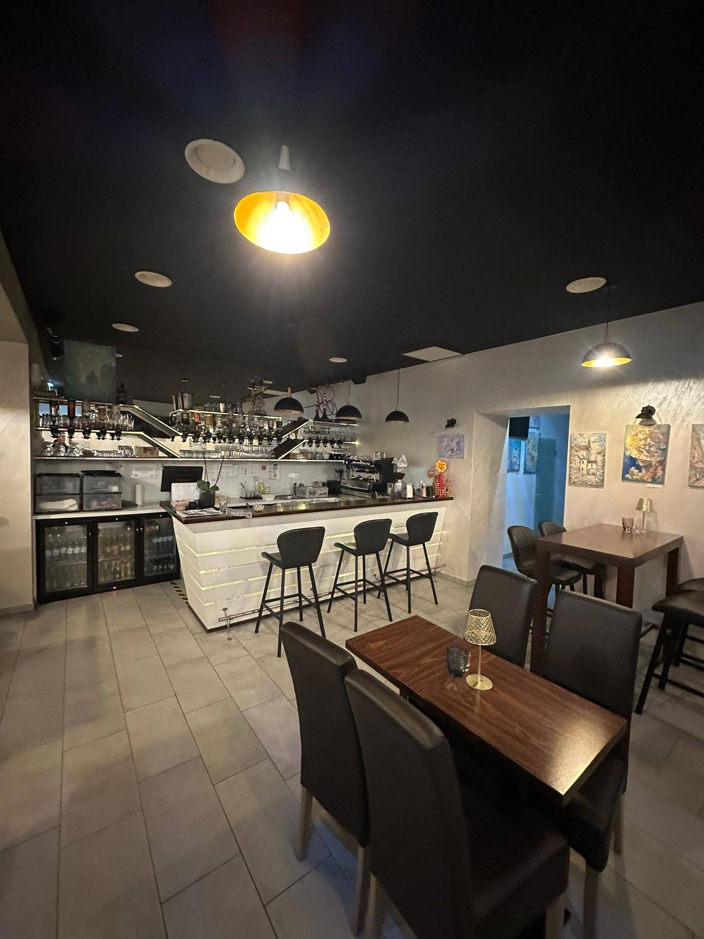 Innenansicht einer Café-Lounge mit Bar, Hockern, Tischen und Stühlen unter einer dunklen Decke.