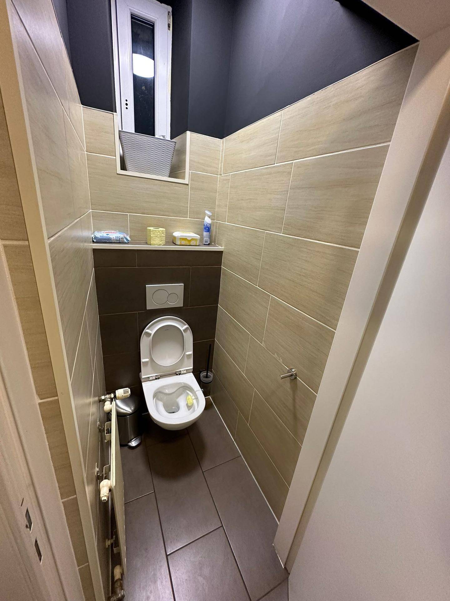 Kleiner Toilettenraum mit wandhängendem WC, beigefarbenen Fliesen und einem Fenster.