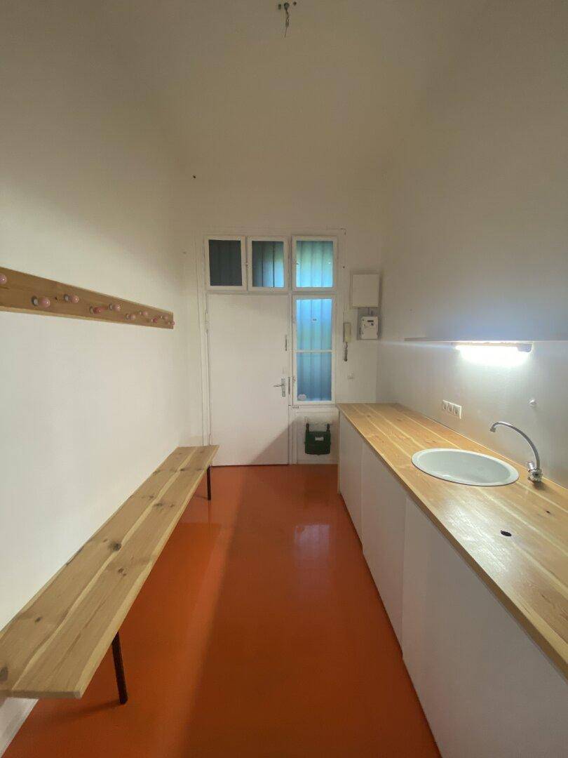 Schmaler Raum mit orangefarbenem Boden, Holztheke mit Spüle und einer Garderobenbank.