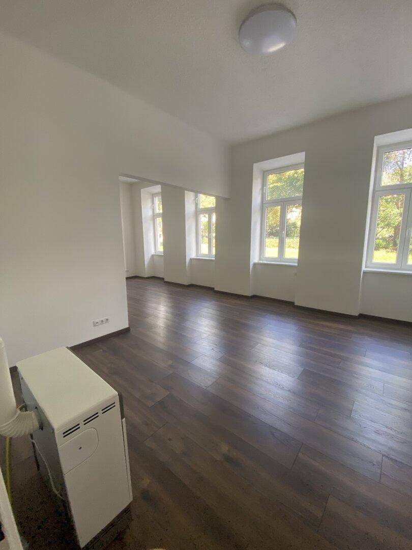 Geräumiger, heller Wohnbereich mit dunklem Holzboden und einem Heizkörper unter dem Fenster.