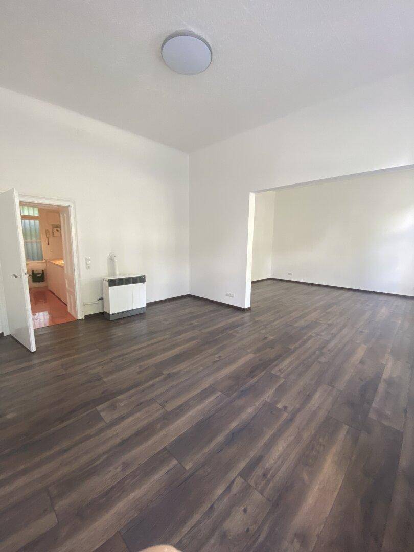 Geräumiger Wohnbereich mit dunklem Holzboden, weiß gestrichenen Wänden und offenem Durchgang zu einem weiteren Raum.