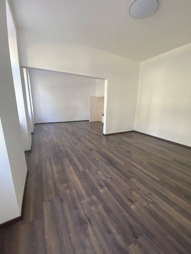 Weitläufiger, heller Raum mit dunklem Holzboden und einem offenen Durchgang zu einem weiteren Bereich.
