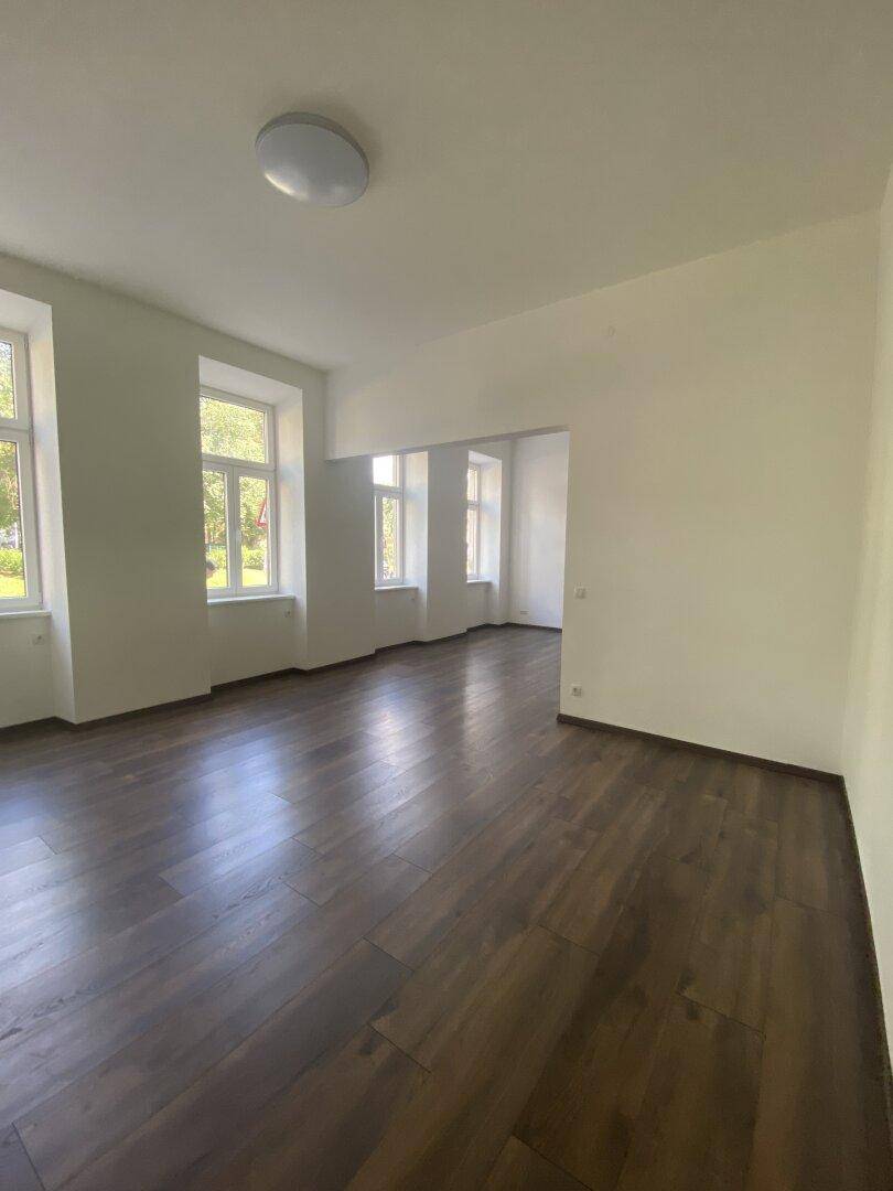 Heller, leerer Raum mit dunklem Holzboden und mehreren Fenstern, die viel Tageslicht hereinlassen.