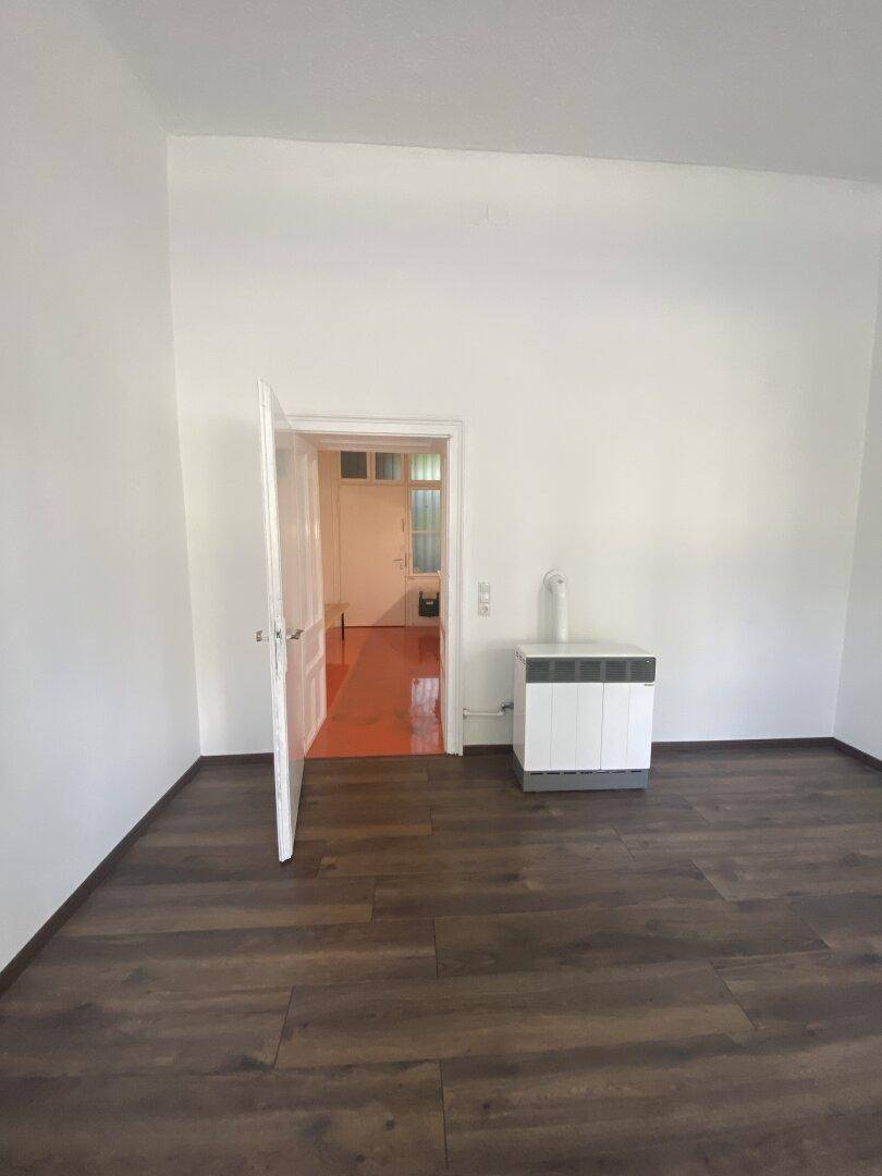 Heller Raum mit dunklem Holzboden und offener Tür, die den Blick in einen orangefarbenen Nebenraum freigibt.