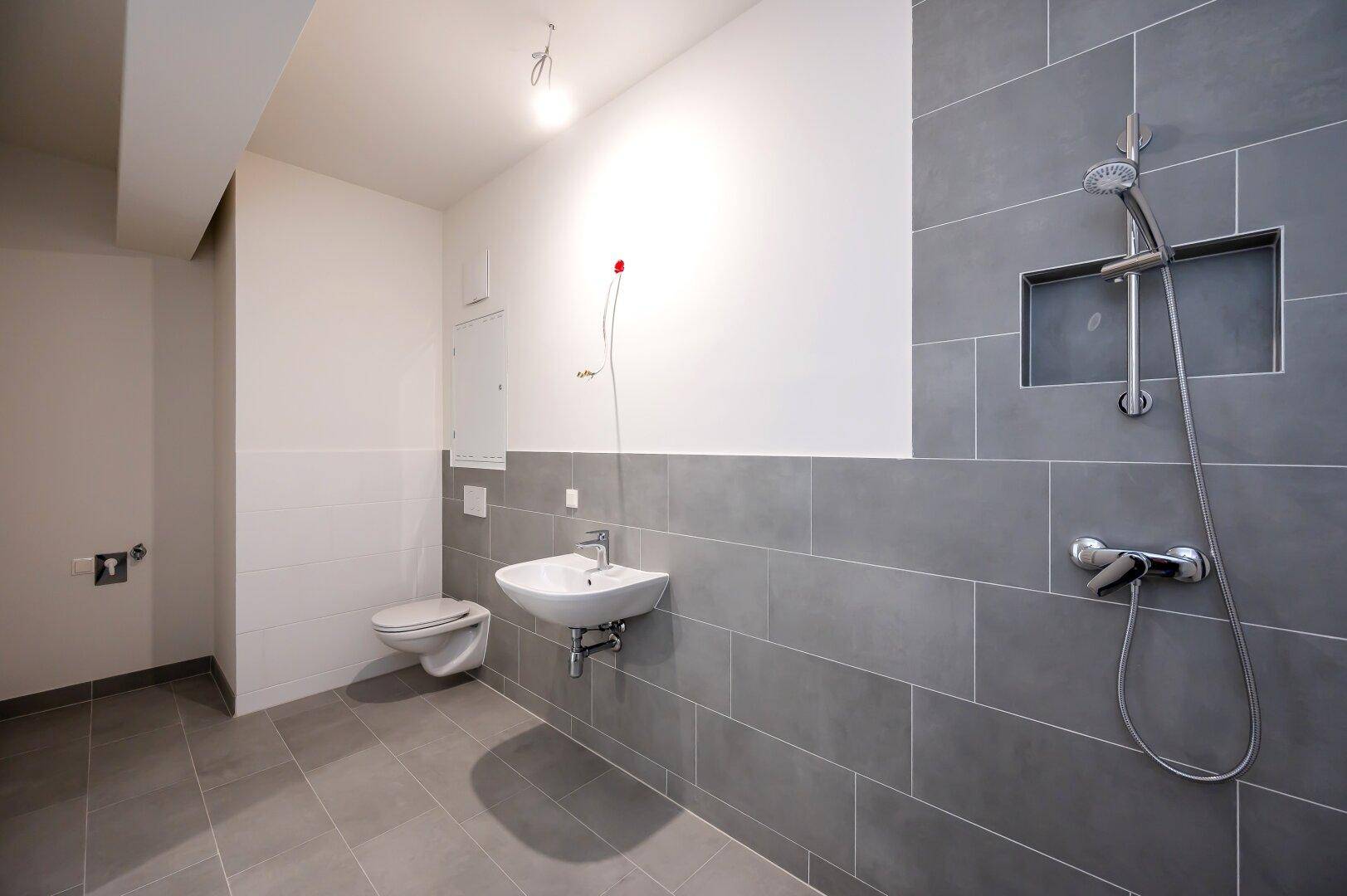 Modernes Badezimmer mit grauen Fliesen, wandhängendem WC und Duschbereich mit Handbrause.