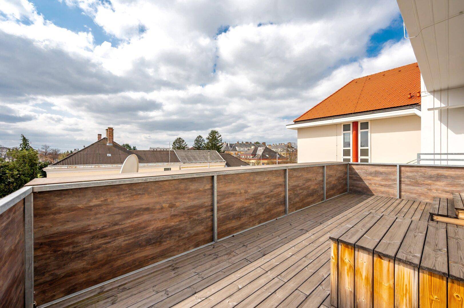 Weitläufige Holzterrasse mit Holzzaun und Blick auf die umliegenden Dächer und Bäume unter blauem Himmel.