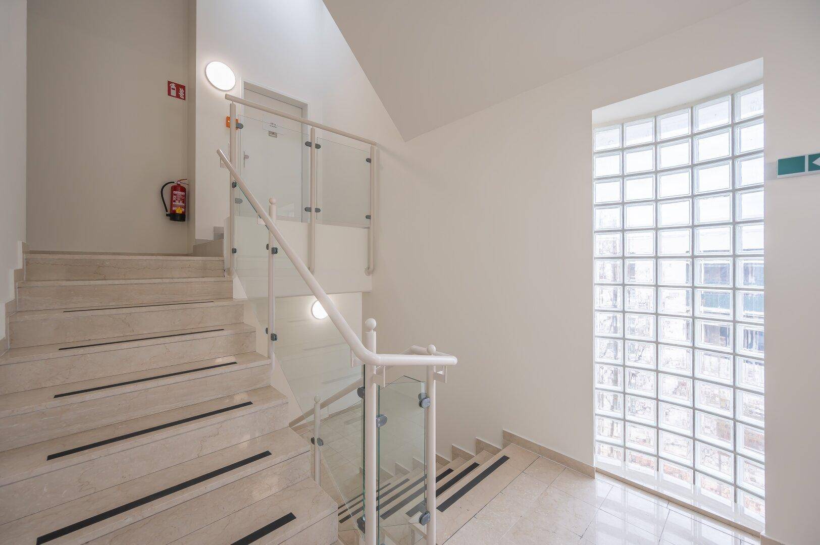Helles Treppenhaus mit Glasbausteinwand, modernem Geländer und Marmorstufen.