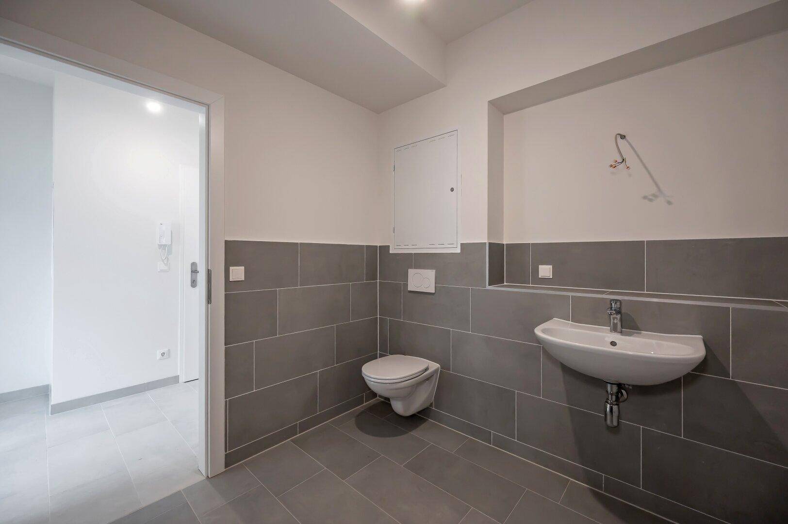 Modernes Gäste-WC mit grauen Fliesen, wandhängendem WC und Waschbecken.