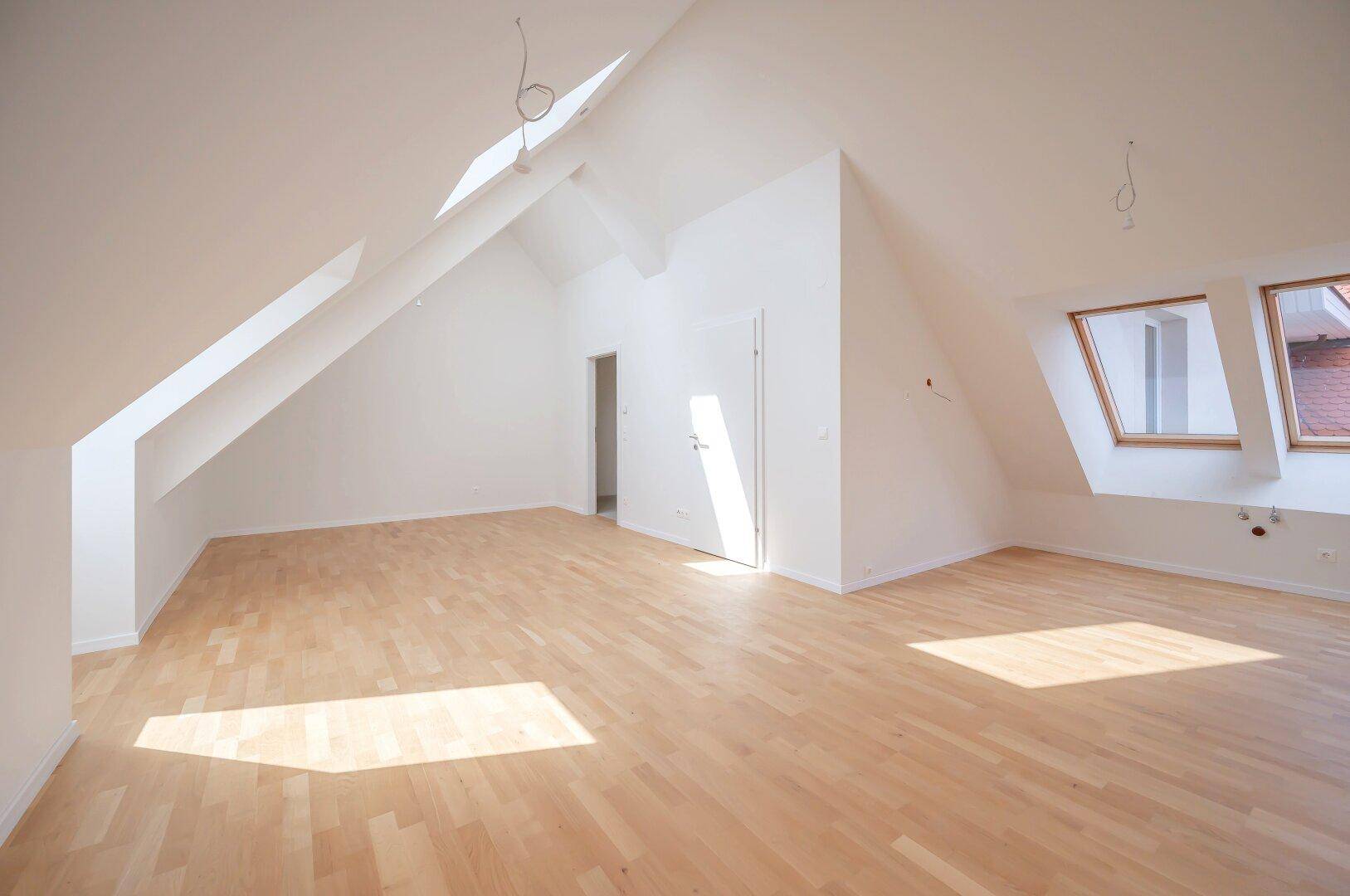 Geräumiger Dachgeschossraum mit Holzparkett, schrägen Wänden und Dachfenstern.