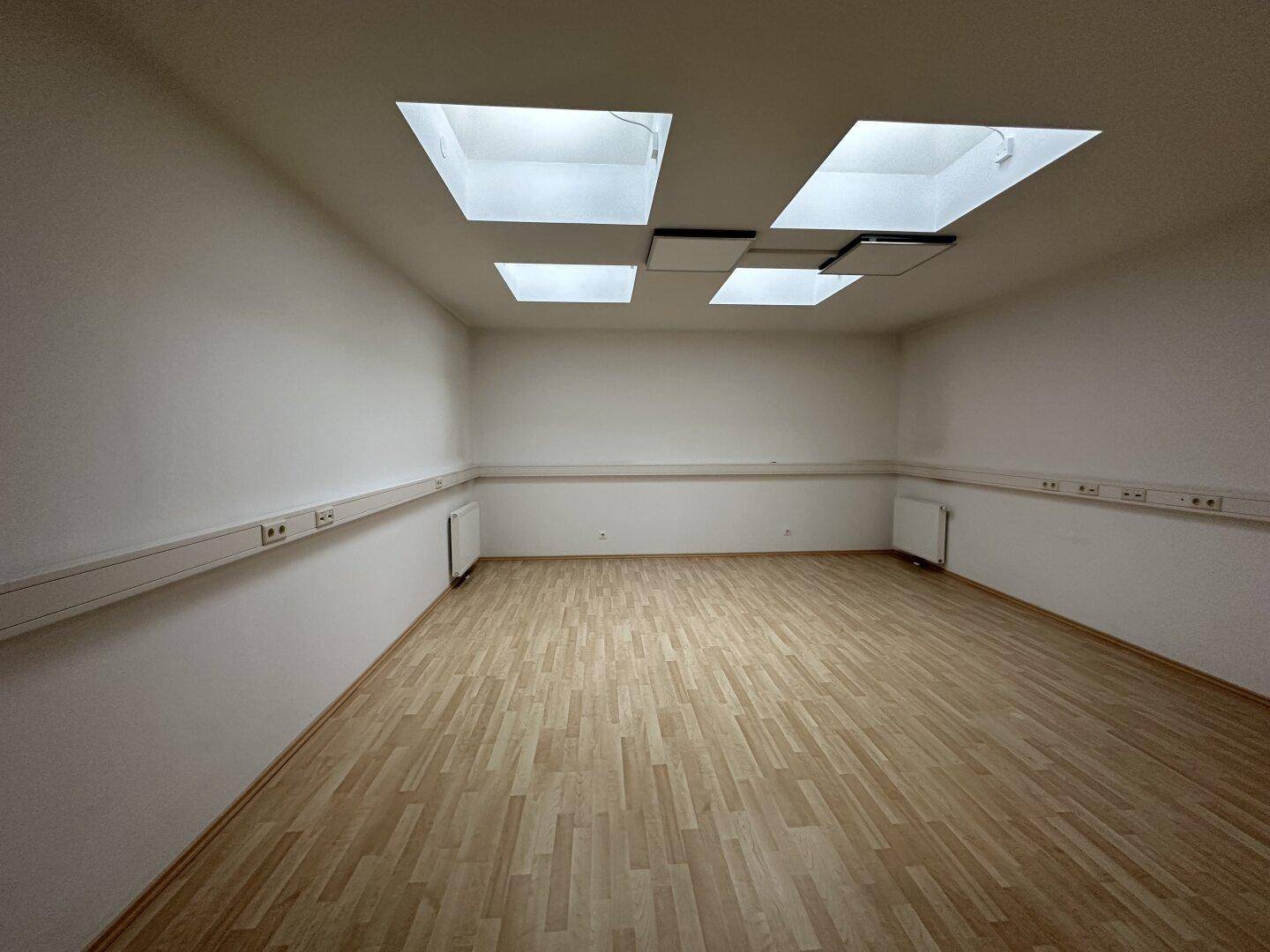 Geräumiger Büroraum mit hellem Holzboden, weißen Wänden und mehreren Dachfenstern für viel Licht.