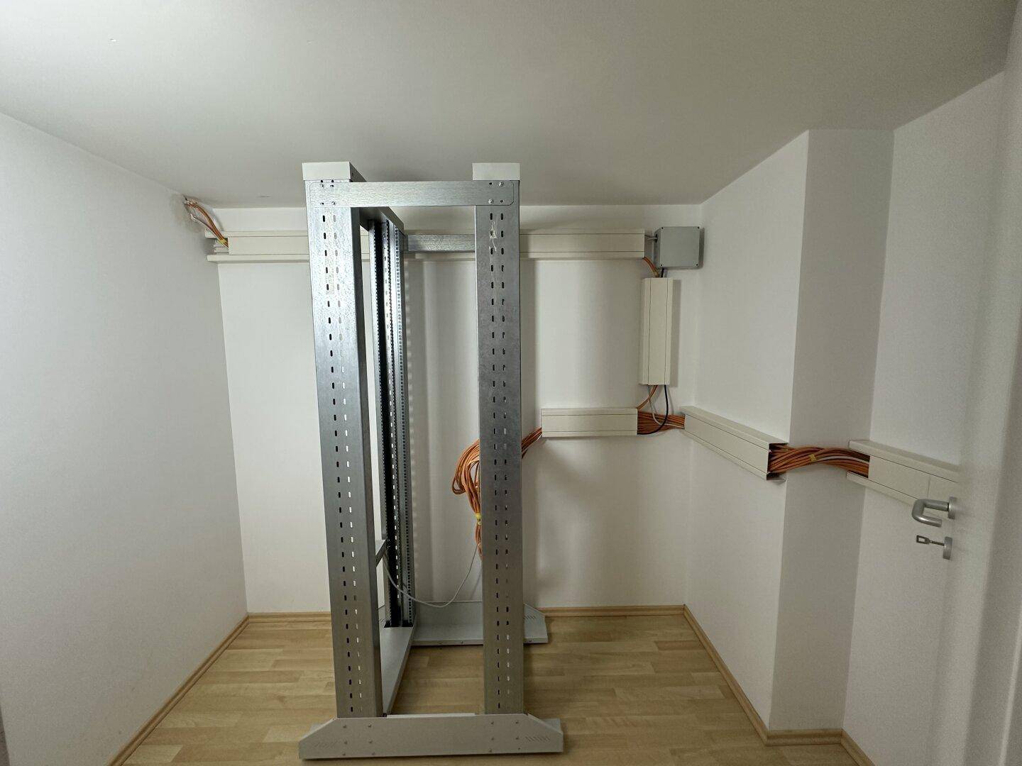 Kleiner Technikraum mit Servergestell und sichtbaren Kabelinstallationen auf Laminatboden.