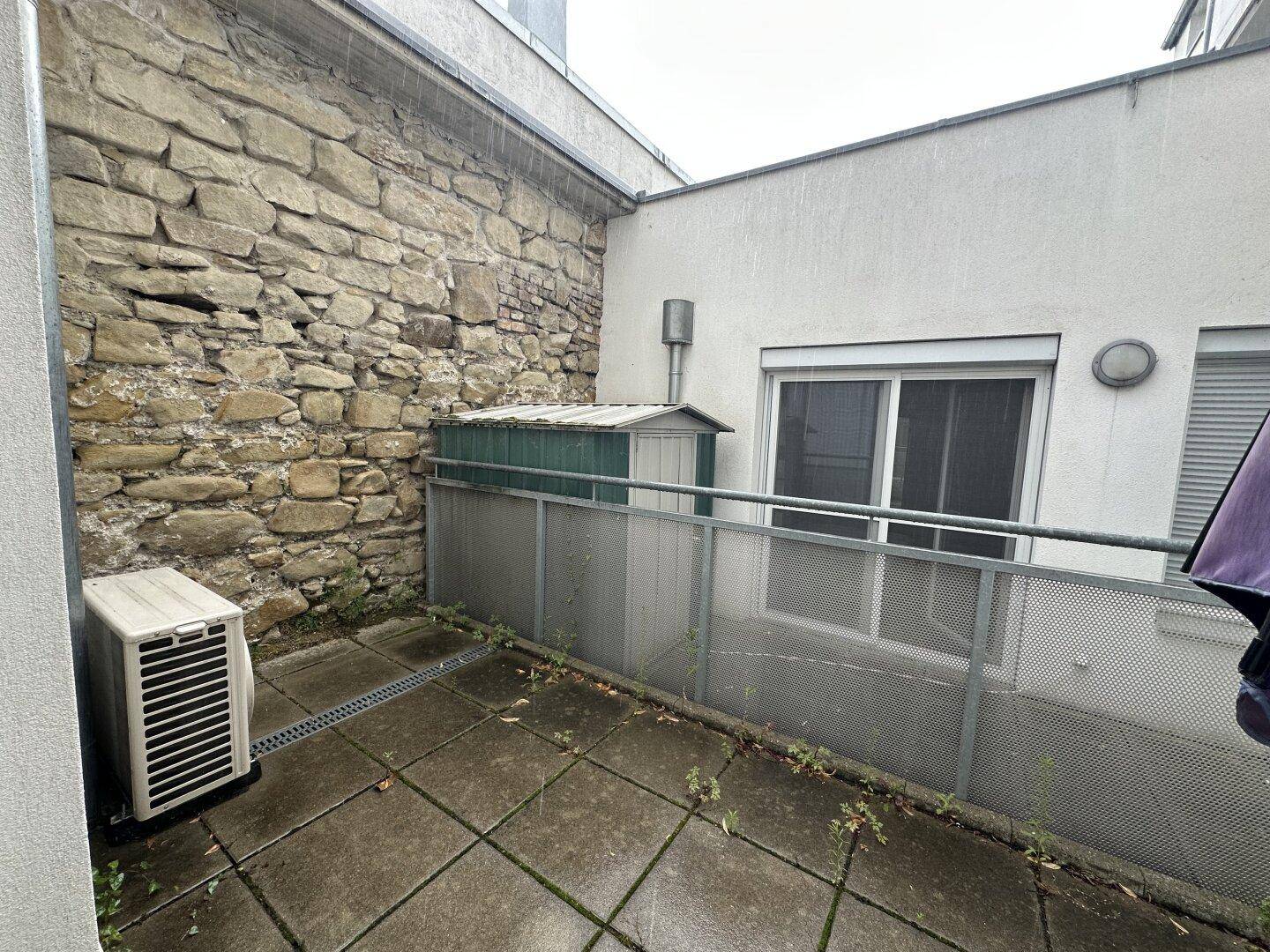 Außenbereich mit Steinmauer, Betonfliesen und einer Klimaanlage, ideal für eine kleine Terrasse.