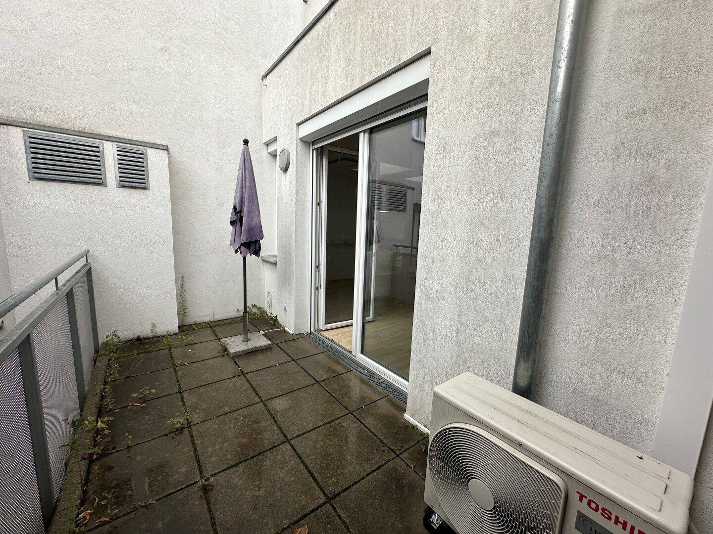 Außenterrasse mit Betonplatten, Sonnenschirm und Klimaanlage, mit Zugang zum Innenbereich.