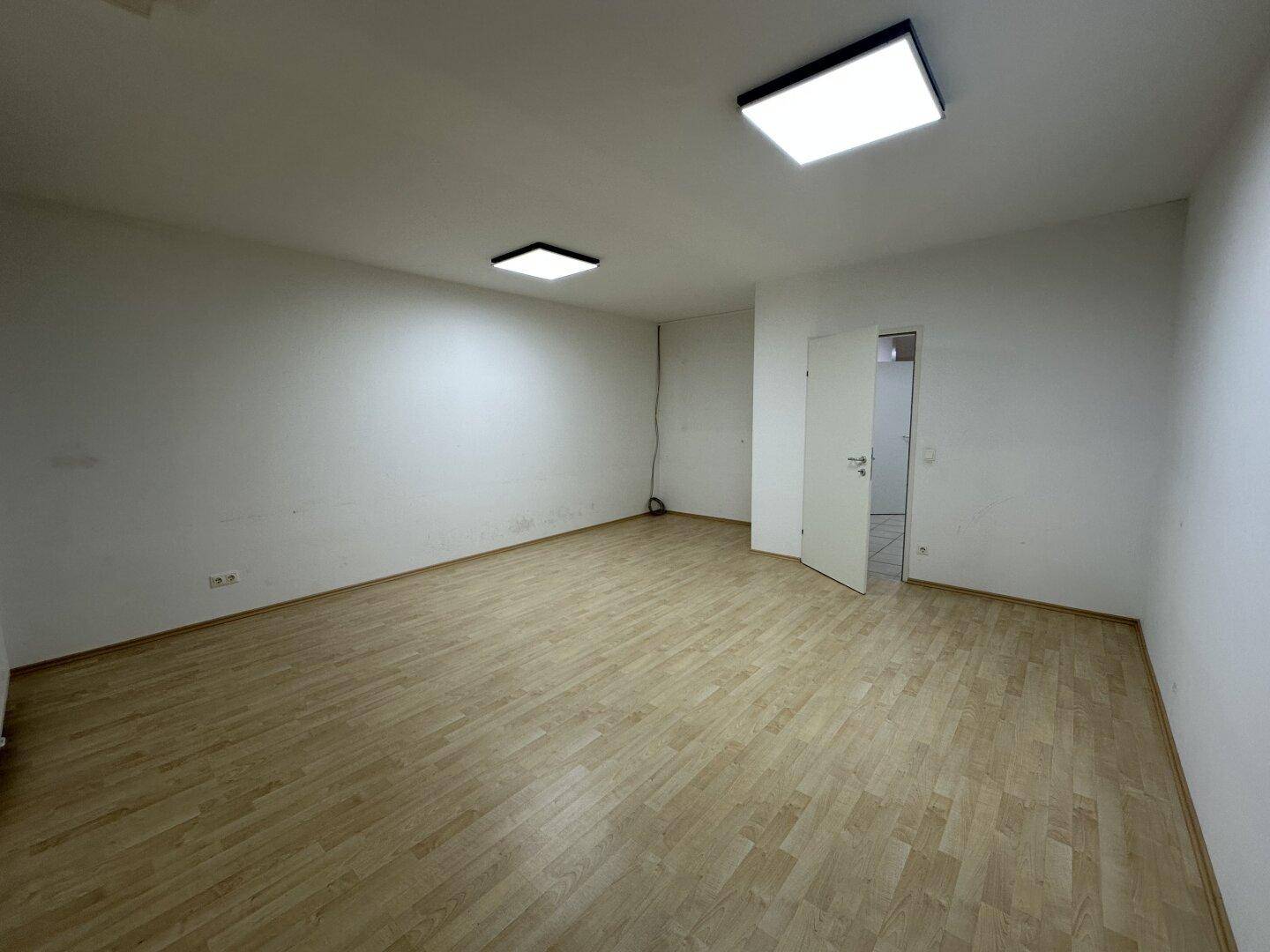 Heller, leerer Büroraum mit Holzboden und zwei Deckenleuchten, der zu einem weiteren Zimmer führt.