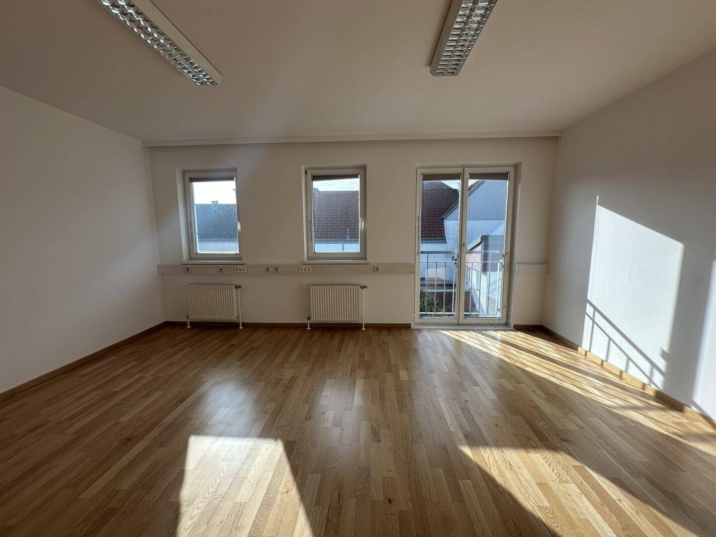 Heller, leerer Raum mit Holzboden, mehreren Fenstern und einem französischen Balkon.