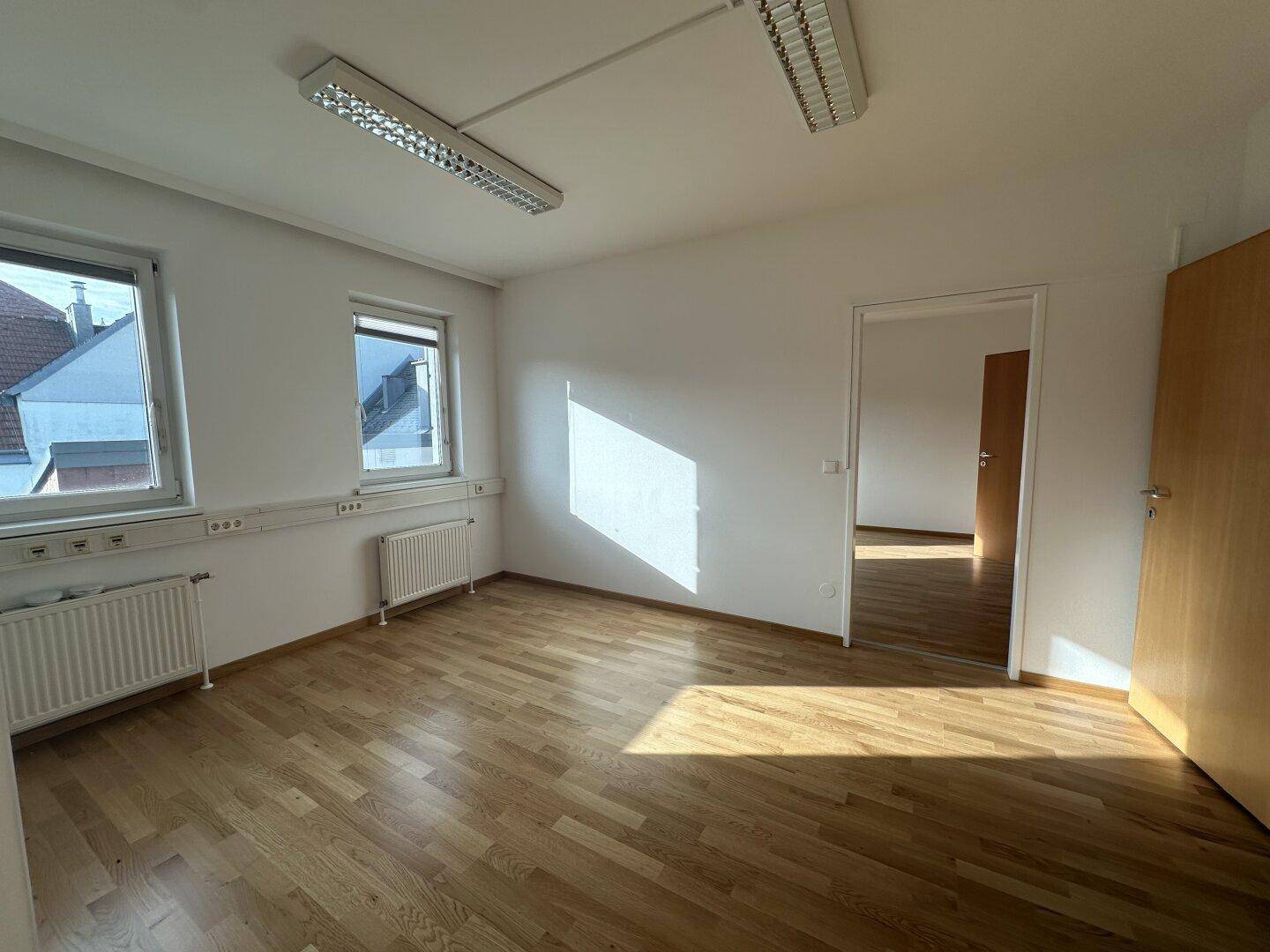 Geräumiger, leerer Raum mit Holzboden, zwei Fenstern und einer Holztür zu einem angrenzenden Bereich.