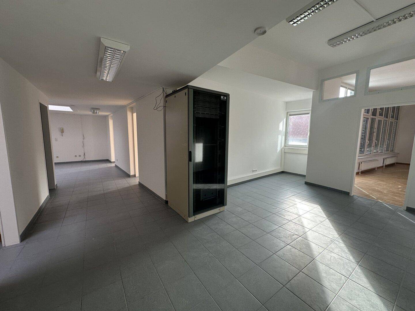 Technischer Raum mit Serverrack, grauen Fliesen und Blick in angrenzende Bürobereiche.