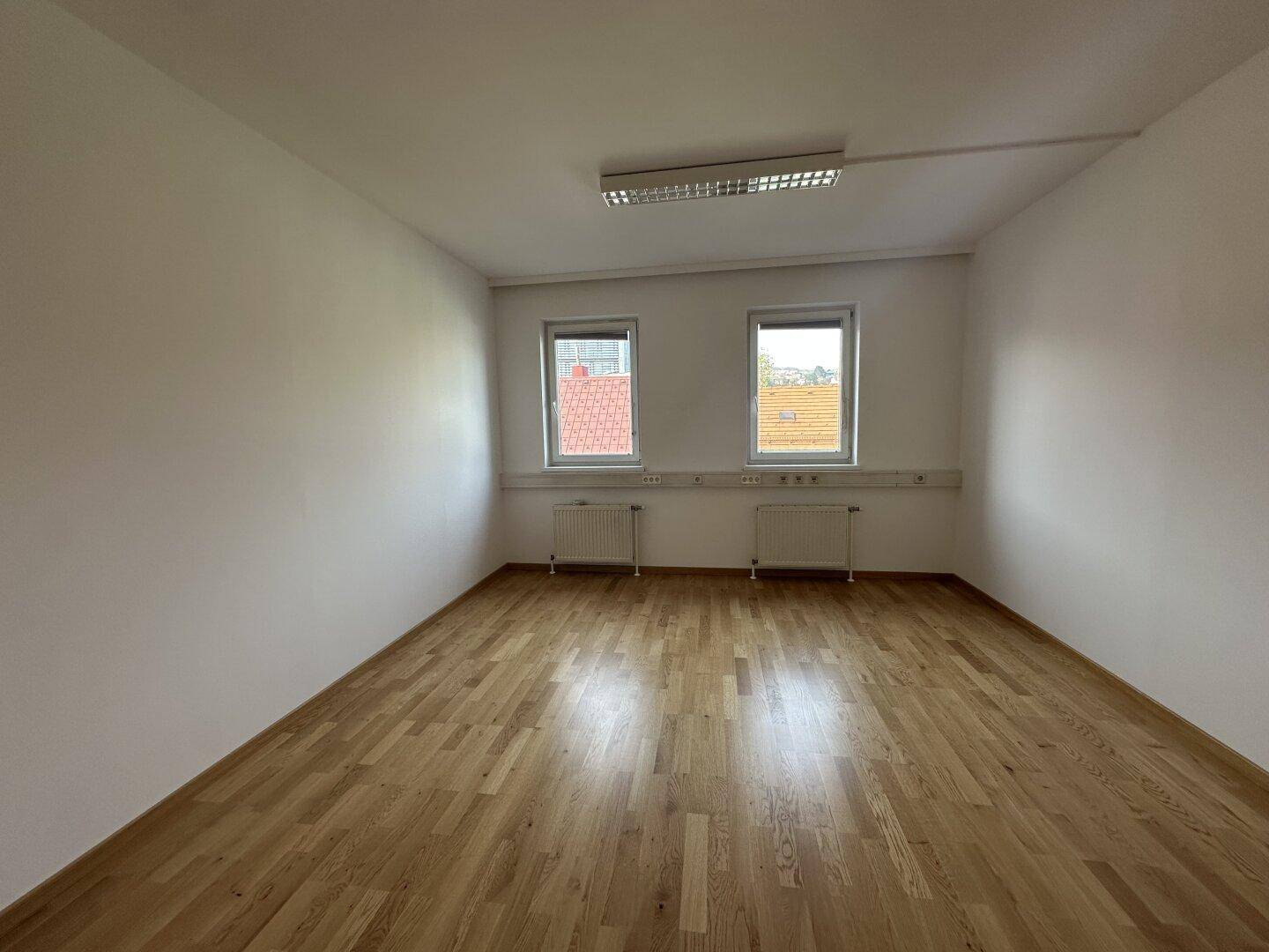 Heller, leerer Büroraum mit Holzboden, zwei Fenstern und Heizkörpern unter den Fenstern.
