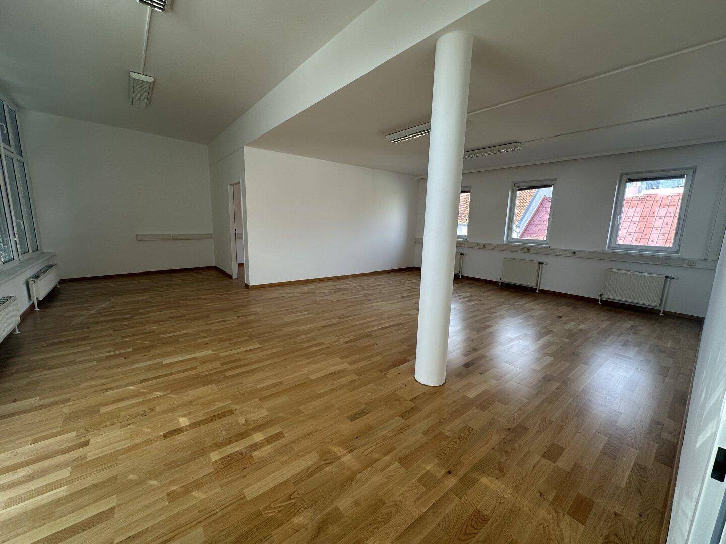 Großer, offener Bürobereich mit Holzboden, Säule und mehreren Fenstern, die viel Licht hereinlassen.
