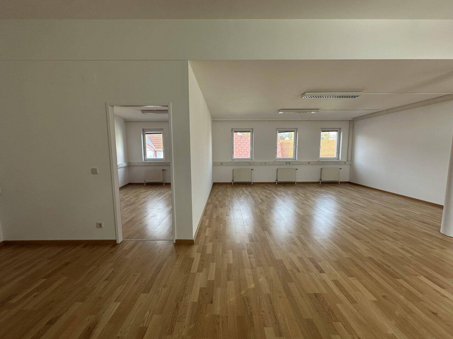 Weitläufiger Bürobereich mit Holzboden und mehreren Fenstern, die den Raum hell und einladend wirken lassen.