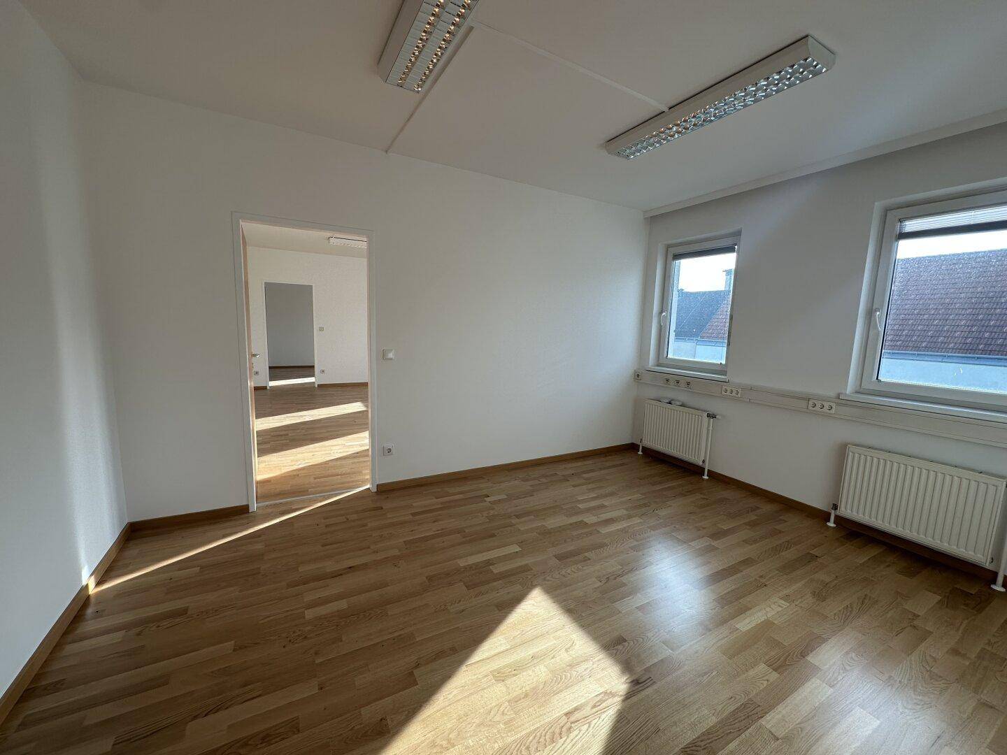 Heller Büroraum mit Holzboden, zwei Fenstern und einer Tür zu einem angrenzenden Zimmer.