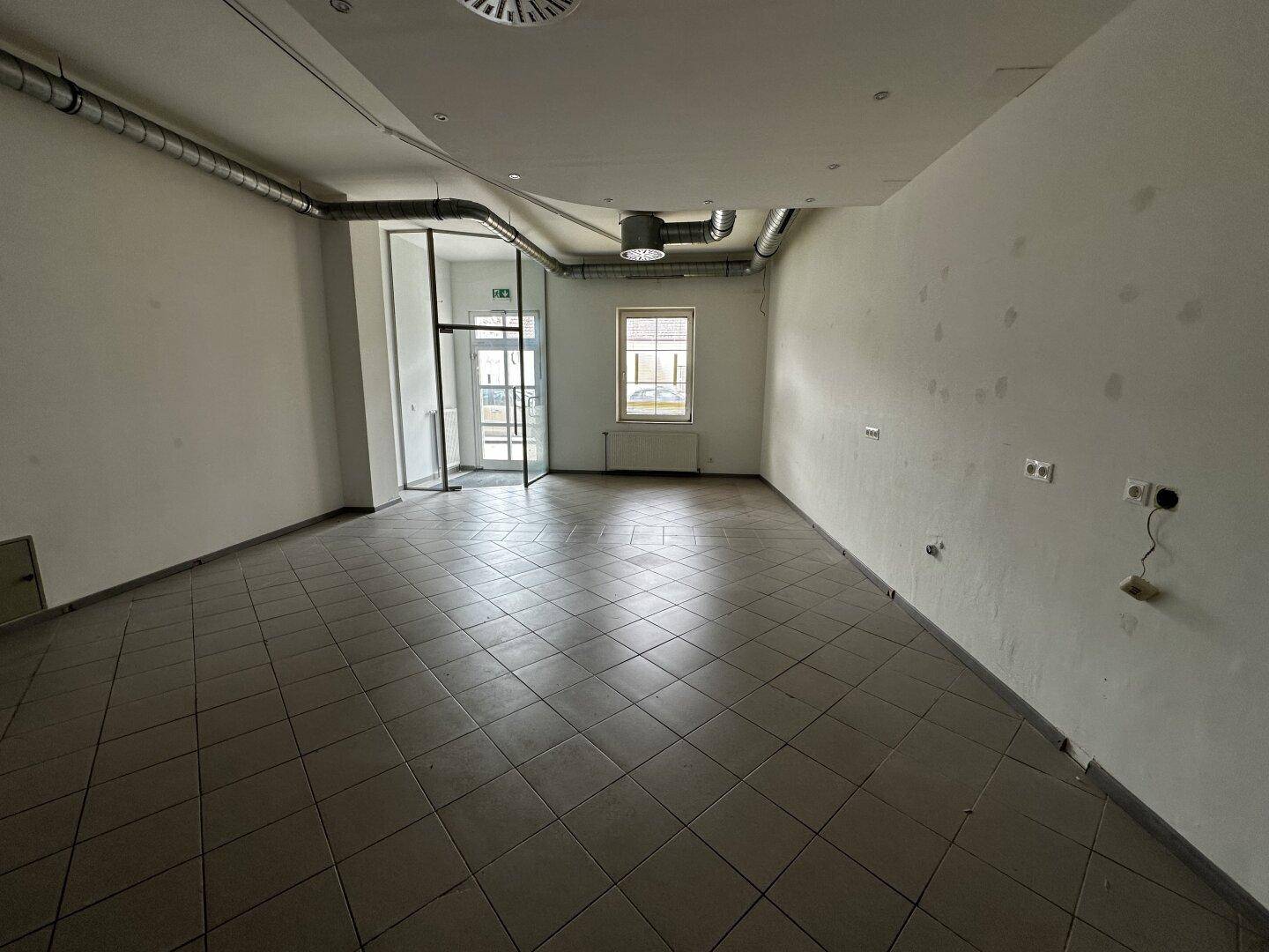 Heller Gewerberaum mit großen Fenstern, Fliesenboden und sichtbaren Lüftungsrohren an der Decke.