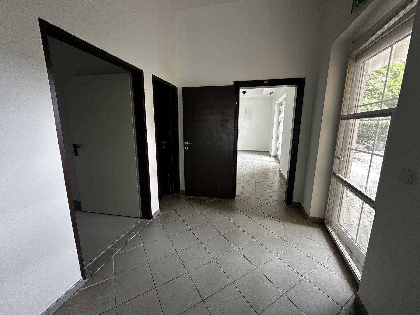 Flur mit Fliesenboden, mehreren Türen und einem großen Fenster, das Tageslicht hereinlässt.