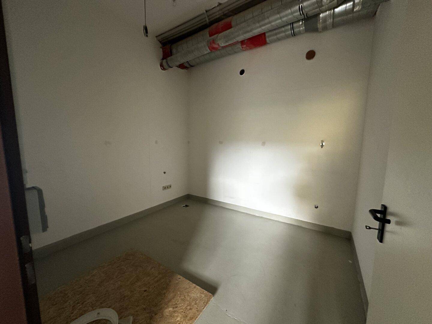 Kleiner Abstellraum mit sichtbaren Lüftungsrohren an der Decke und einfachem Bodenbelag.