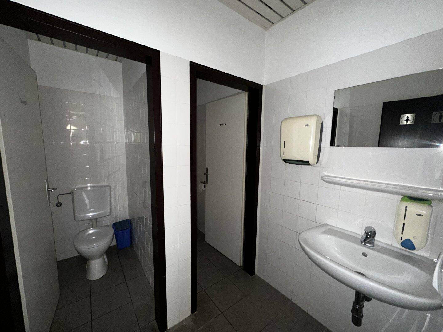 Toilettenbereich mit Waschbecken, Spiegel und zwei separaten Kabinen, gefliest in Weiß und Grau.