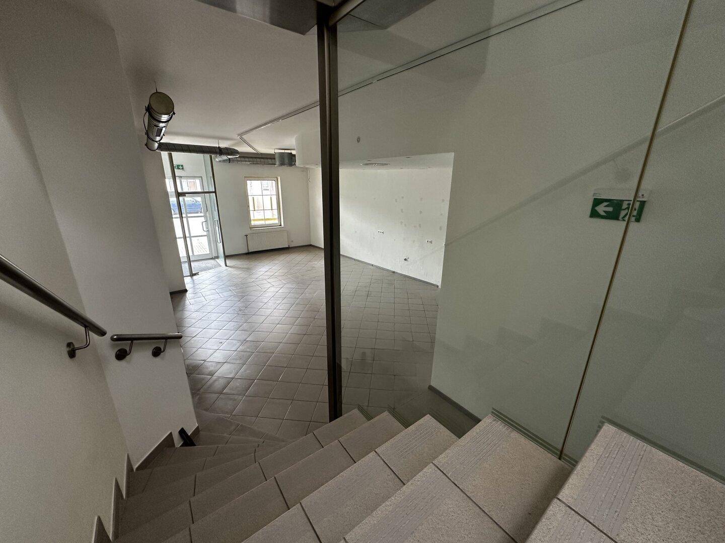 Treppenhaus mit Glasgeländer und Blick in den hellen Gewerberaum im Erdgeschoss.