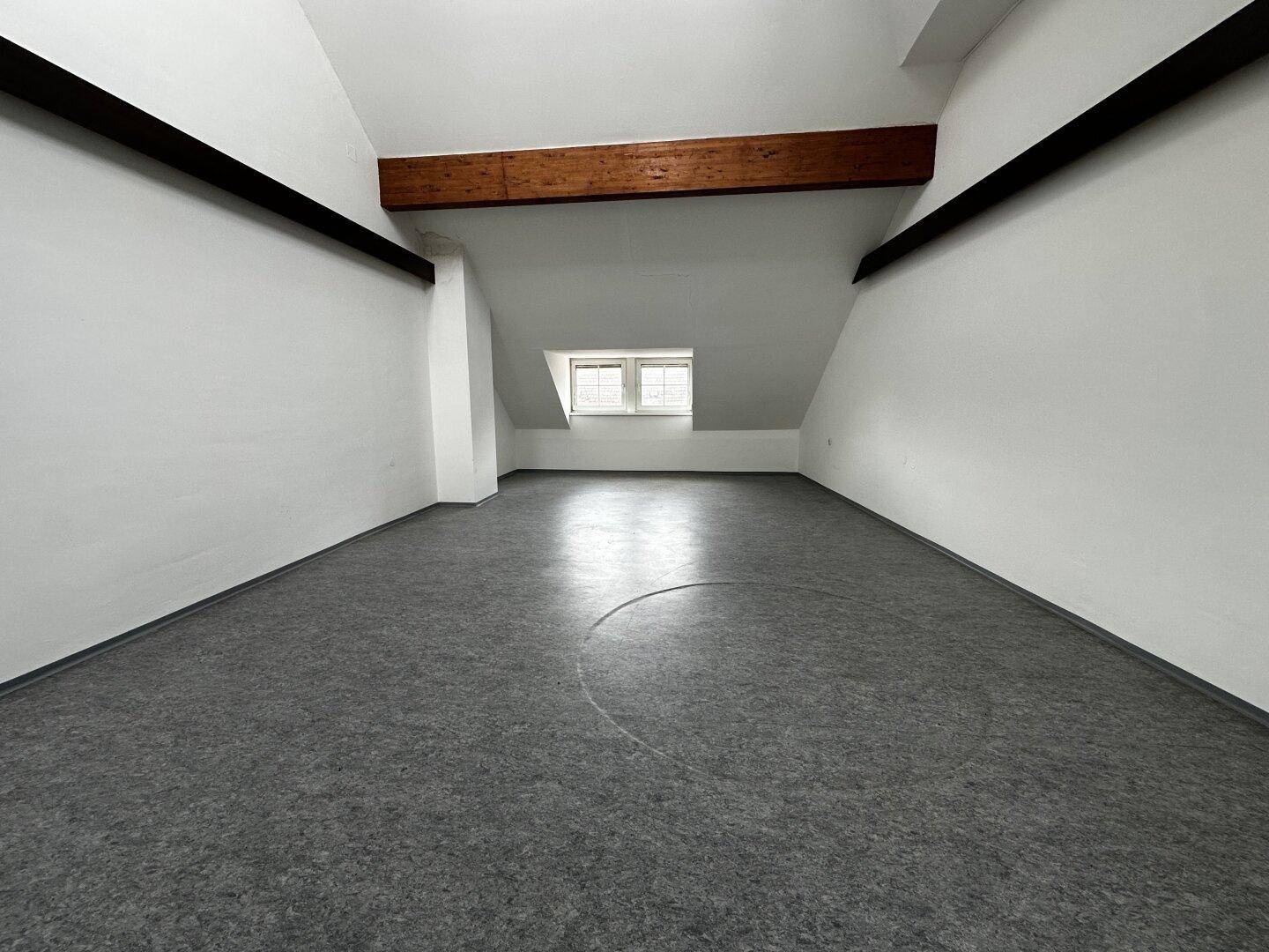 Langer Raum im Dachgeschoss mit schrägen Wänden, Holzbalken und einem kleinen Fenster.