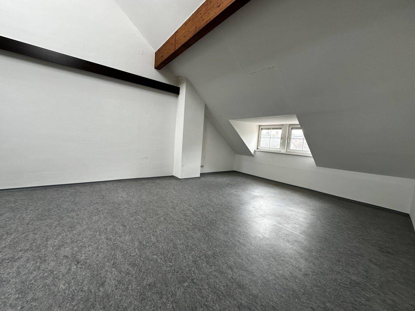 Heller Raum im Dachgeschoss mit schrägen Wänden, Holzbalken und zwei Fenstern.