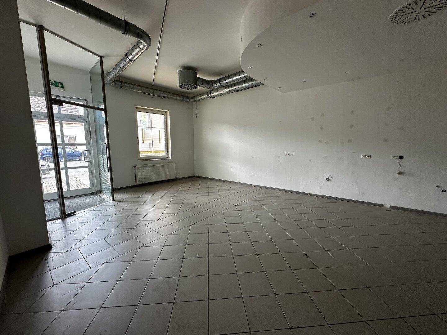 Heller Gewerberaum mit Fliesenboden, großen Fenstern und sichtbaren Lüftungsrohren an der Decke.
