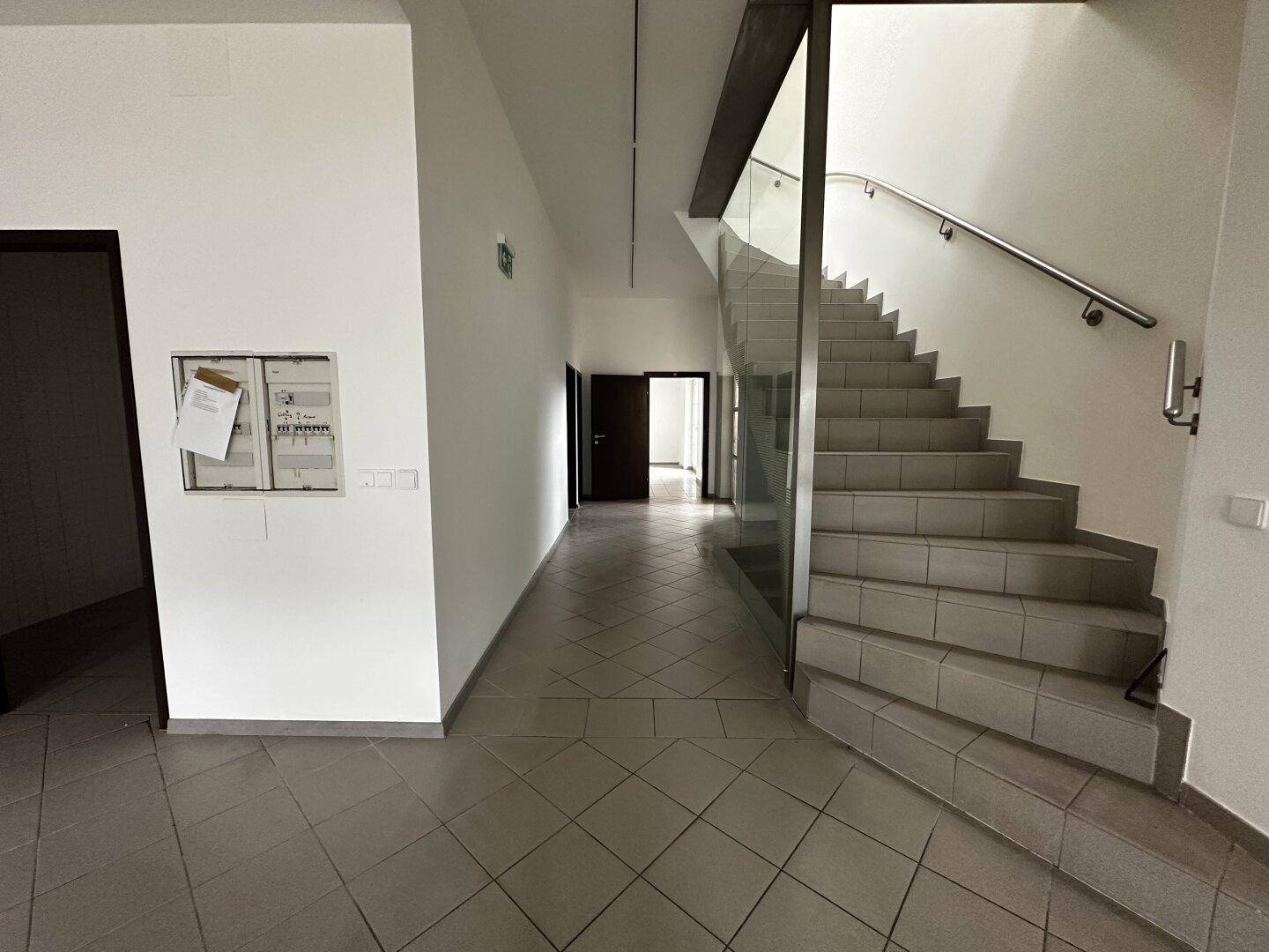 Langer Flur mit Fliesenboden, Treppe mit Glasgeländer und Zugang zu weiteren Räumen.