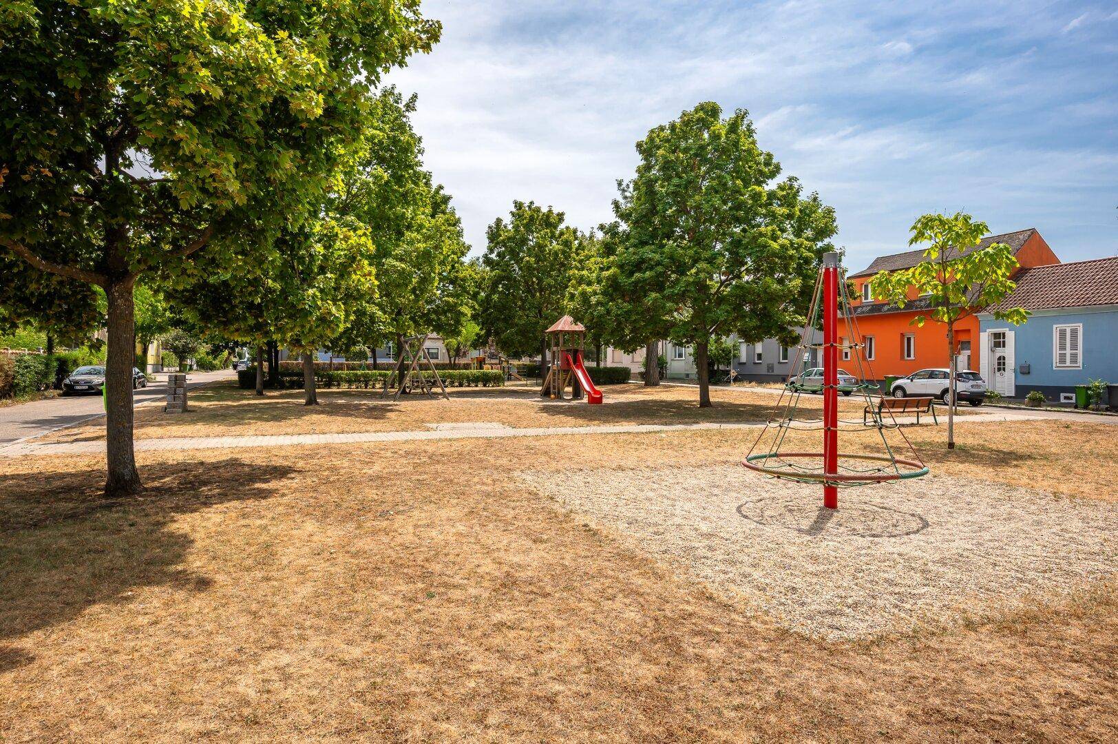 Öffentlicher Spielplatz mit Bäumen, Rasenflächen und verschiedenen Spielgeräten für Kinder.