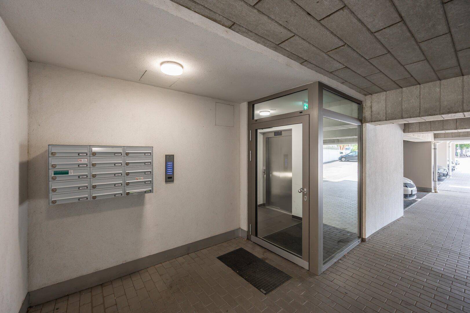 Eingangsbereich des Gebäudes mit Briefkästen, Gegensprechanlage und modernem Glasaufzug.