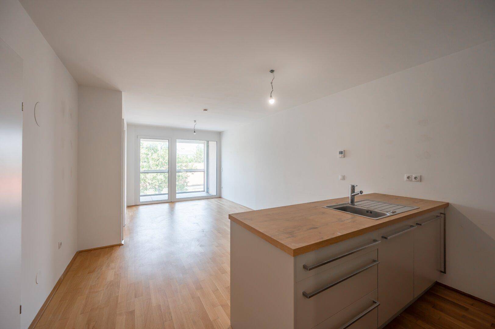 Heller Wohnbereich mit Parkettboden und großen Fenstern, die viel Tageslicht hereinlassen und Zugang zum Balkon bieten.