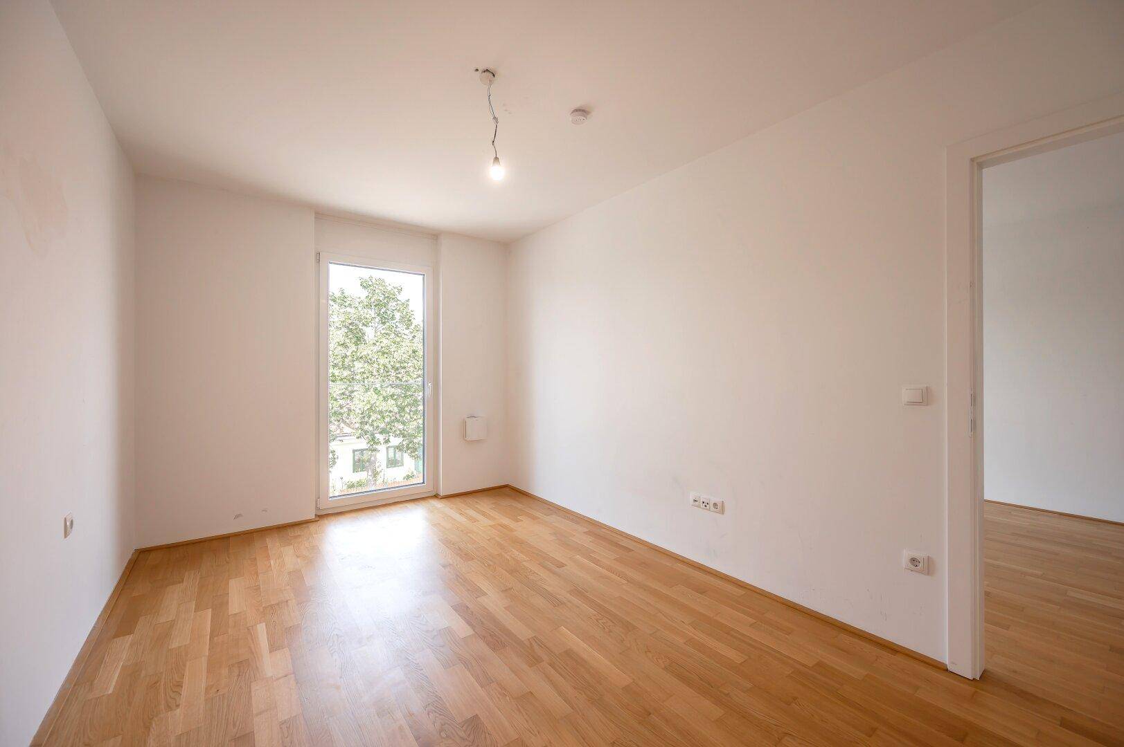 Helles Schlafzimmer mit Parkettboden und einem großen Fenster, das eine angenehme Atmosphäre schafft.
