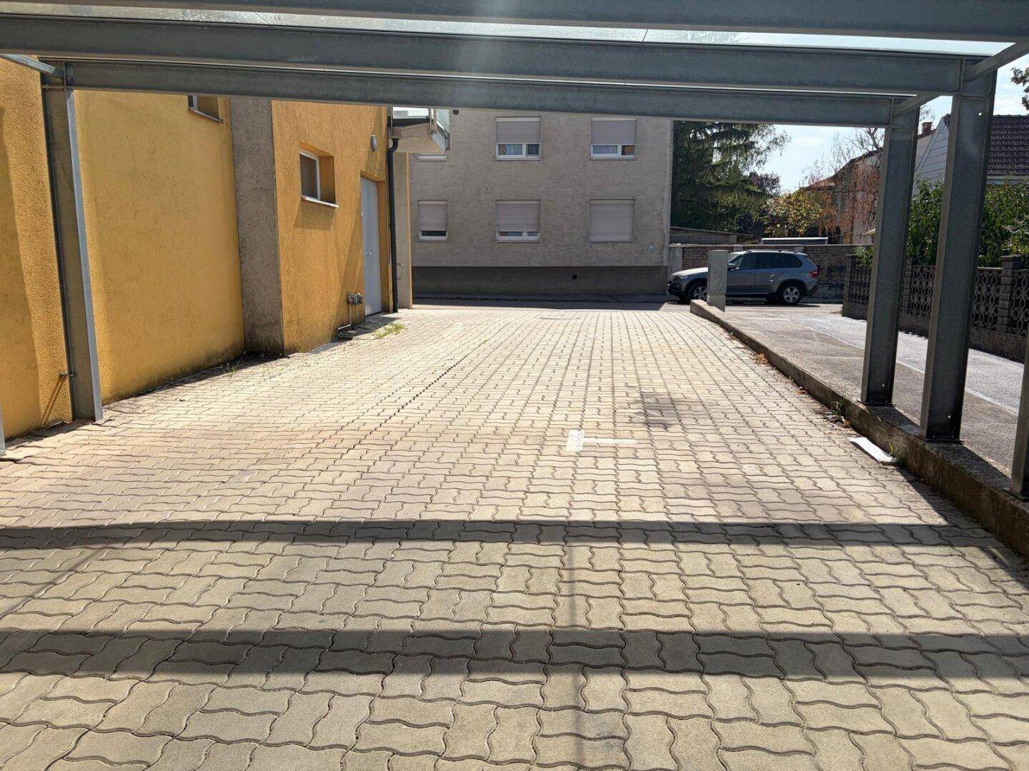 Weitläufiger gepflasterter Bereich unter einem Carport, mit angrenzendem Gebäude und Blick auf die Umgebung.