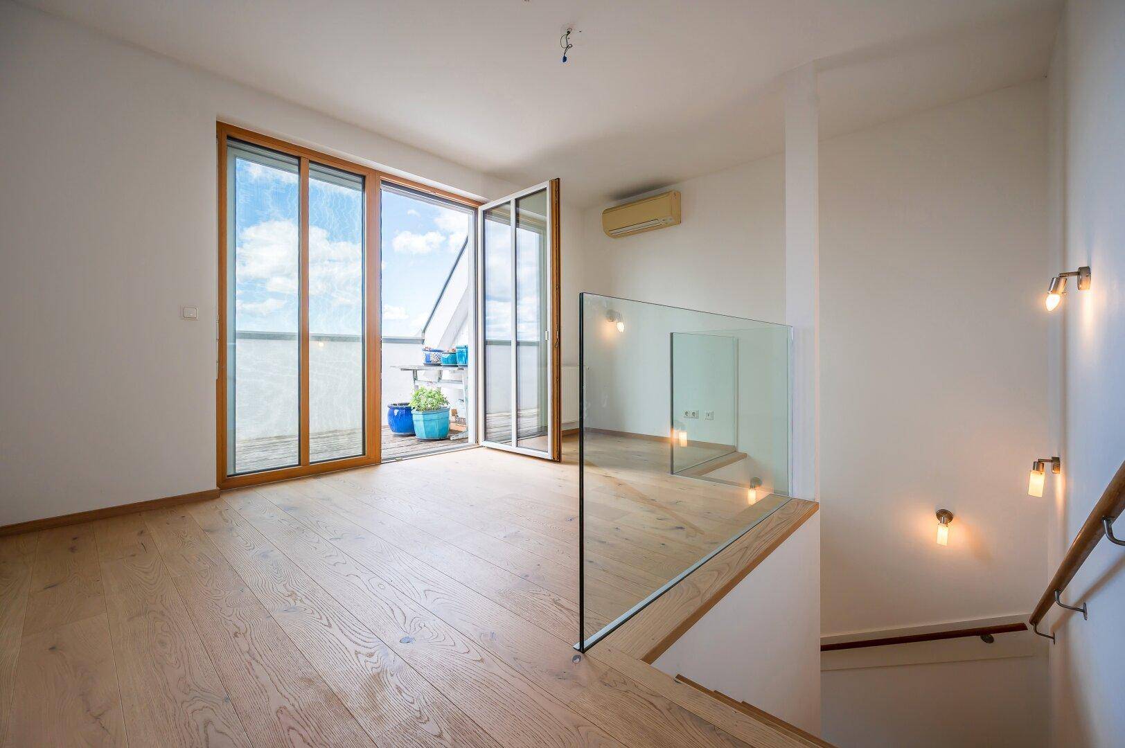 Heller Wohnbereich im Obergeschoss mit Glasgeländer und Zugang zu einem sonnigen Balkon.