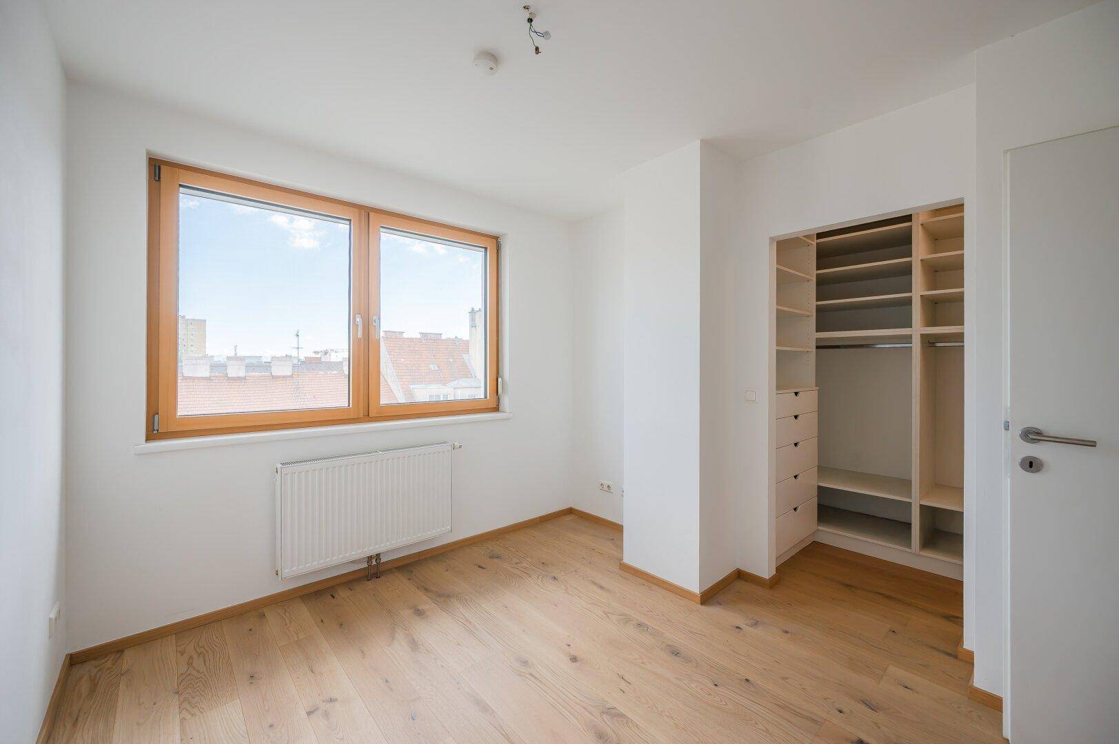 Helles Schlafzimmer mit großem Fenster, Holzboden und integriertem Kleiderschrank.