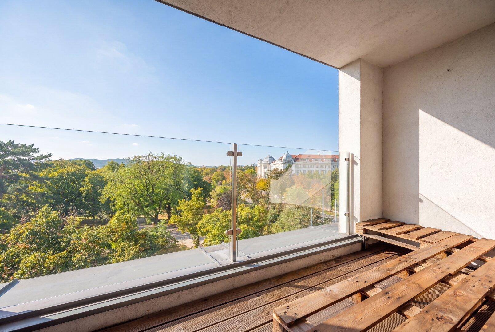 Sonniger Balkon mit Glasgeländer, Holzboden und weitem Blick ins Grüne.