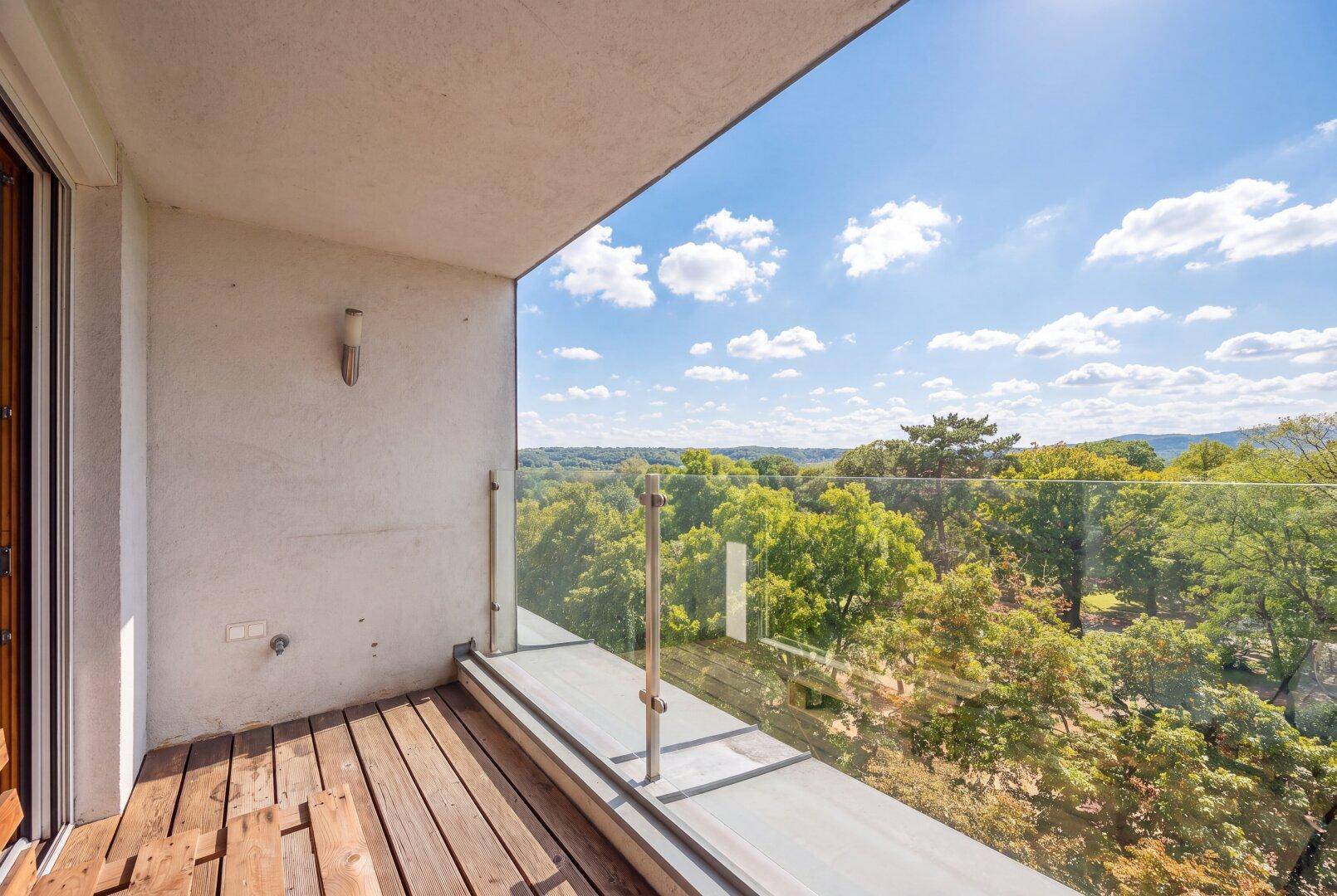 Heller Balkon mit Holzbelag, Glasgeländer und Panoramablick über die Baumwipfel.